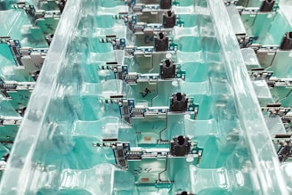 A collection of electronic circuit boards organized neatly in transparent plastic trays. The boards are lined in rows, displaying components, connectors, and intricate circuitry on a teal background.