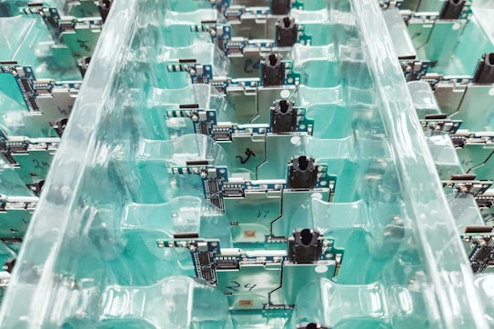 A collection of electronic circuit boards organized neatly in transparent plastic trays. The boards are lined in rows, displaying components, connectors, and intricate circuitry on a teal background.