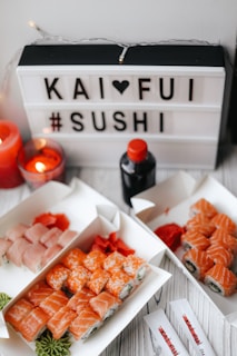 A lightbox with letters spelling out 'KAI ❤️ FUI #SUSHI' is surrounded by sushi platters and candles. There are various sushi rolls in the trays, with colors ranging from salmon pink to orange. A bottle of soy sauce is also visible on the table.