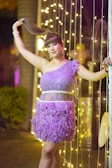 A person stands confidently in a glamorous purple dress adorned with sparkles and feathers. They are holding their hair up with one hand, while surrounded by vertical strings of warm, glowing lights. The background includes tall plants and blurred lighting, suggesting a festive or celebratory environment.