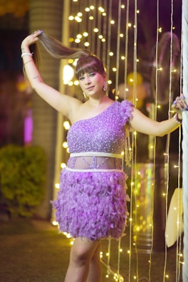 A person stands confidently in a glamorous purple dress adorned with sparkles and feathers. They are holding their hair up with one hand, while surrounded by vertical strings of warm, glowing lights. The background includes tall plants and blurred lighting, suggesting a festive or celebratory environment.