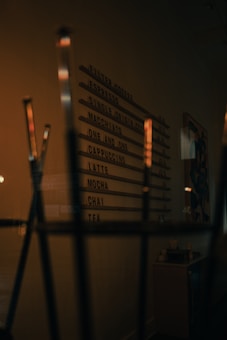 A dimly lit coffee shop menu board lists various beverages such as espresso, cappuccino, and chai. The surrounding environment has a cozy, warm ambiance with elements like a counter and decor visible in the background.