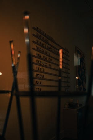A dimly lit coffee shop menu board lists various beverages such as espresso, cappuccino, and chai. The surrounding environment has a cozy, warm ambiance with elements like a counter and decor visible in the background.