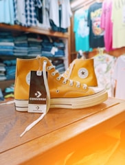 A pair of yellow high-top Converse sneakers displayed on a wooden surface. The sneakers have white laces and a tag attached to one of them. In the background, there are various clothing items such as folded jeans and hanging shirts in different colors.
