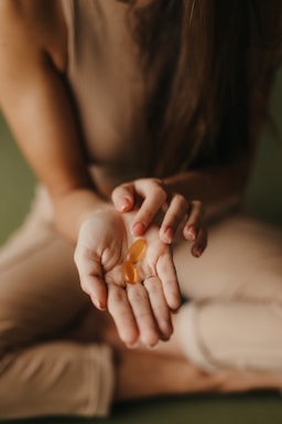 A person wearing a brown sleeveless top and light pants is sitting cross-legged on a soft surface, holding two oval-shaped orange capsules in their open palm.