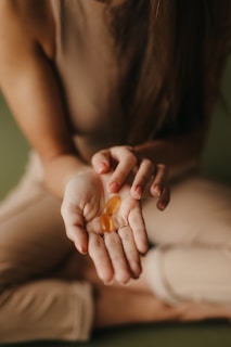 A person wearing a brown sleeveless top and light pants is sitting cross-legged on a soft surface, holding two oval-shaped orange capsules in their open palm.
