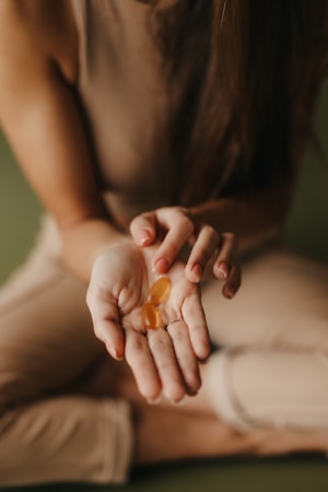 A person wearing a brown sleeveless top and light pants is sitting cross-legged on a soft surface, holding two oval-shaped orange capsules in their open palm.