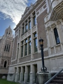 A historical building with ornate stone architecture, featuring large windows and intricate carvings. The structure has a classic, academic or institutional appearance, with a tall tower and a decorative lamppost in the foreground. The sky is partly cloudy, with sunlight peeking through.