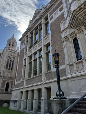 A historical building with ornate stone architecture, featuring large windows and intricate carvings. The structure has a classic, academic or institutional appearance, with a tall tower and a decorative lamppost in the foreground. The sky is partly cloudy, with sunlight peeking through.