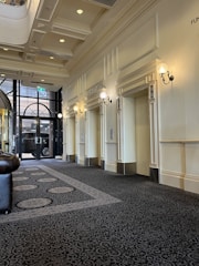 A sophisticated hallway featuring guest lifts with elegant wall sconces. The ceiling is adorned with recessed lighting and decorative molding. The floor is covered with a detailed patterned carpet. Large glass doors at the end of the hallway show glimpses of an exterior view.