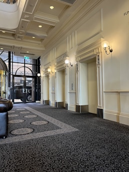 A sophisticated hallway featuring guest lifts with elegant wall sconces. The ceiling is adorned with recessed lighting and decorative molding. The floor is covered with a detailed patterned carpet. Large glass doors at the end of the hallway show glimpses of an exterior view.