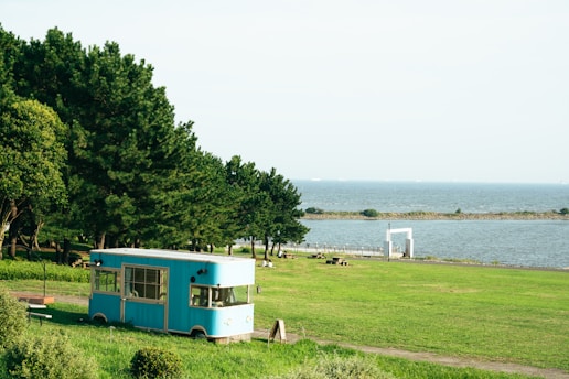 A turquoise food truck is parked on a lush green lawn near a scenic coastline. The backdrop includes a large body of water and a line of trees providing shade. A few benches and tables are scattered across the grass, and a small white structure is visible in the distance.