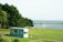 A turquoise food truck is parked on a lush green lawn near a scenic coastline. The backdrop includes a large body of water and a line of trees providing shade. A few benches and tables are scattered across the grass, and a small white structure is visible in the distance.