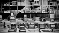 A collection of vintage computers and monitors displayed on shelves and desks in a museum or exhibition setting. The devices vary in size and design, with some displaying graphics and interfaces typical of older computer systems. The arrangement is neat, with some additional tech memorabilia and decorative pieces enhancing the retro atmosphere.