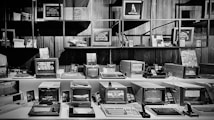 A collection of vintage computers and monitors displayed on shelves and desks in a museum or exhibition setting. The devices vary in size and design, with some displaying graphics and interfaces typical of older computer systems. The arrangement is neat, with some additional tech memorabilia and decorative pieces enhancing the retro atmosphere.