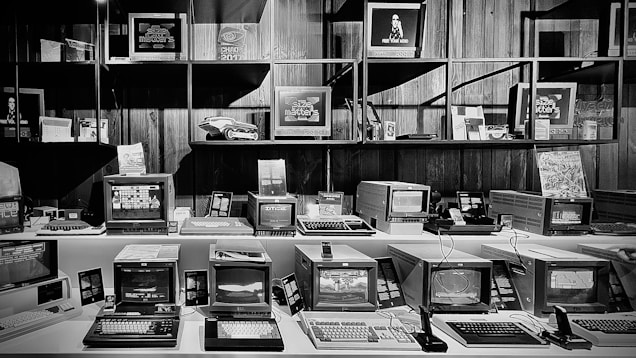 A collection of vintage computers and monitors displayed on shelves and desks in a museum or exhibition setting. The devices vary in size and design, with some displaying graphics and interfaces typical of older computer systems. The arrangement is neat, with some additional tech memorabilia and decorative pieces enhancing the retro atmosphere.