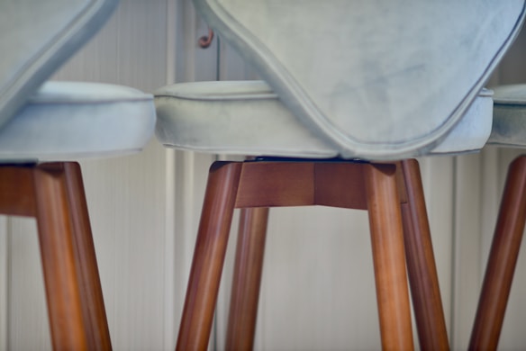 A close-up view of cushioned bar stools with light grey upholstery and wooden legs, set against a softly lit interior background.