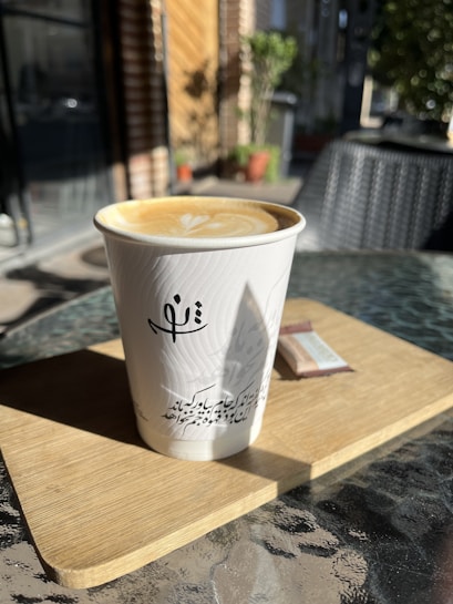 A cup of cappuccino sits on a wooden board, placed on a glass table. Sunlight casts shadows, creating a cozy outdoor caf&eacute; ambiance. In the blurred background, potted plants and wicker seating can be seen. The cup features a latte art design and has writing in a language resembling Arabic script.