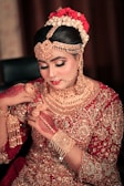 A woman dressed in traditional bridal attire adorned with intricate gold jewelry covering her neck, ears, and forehead. Her hair is styled elegantly with flowers, and her hands are decorated with henna designs. The outfit is richly embroidered with gold embellishments on a red fabric.