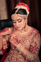 A woman dressed in traditional bridal attire adorned with intricate gold jewelry covering her neck, ears, and forehead. Her hair is styled elegantly with flowers, and her hands are decorated with henna designs. The outfit is richly embroidered with gold embellishments on a red fabric.