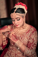 A woman dressed in traditional bridal attire adorned with intricate gold jewelry covering her neck, ears, and forehead. Her hair is styled elegantly with flowers, and her hands are decorated with henna designs. The outfit is richly embroidered with gold embellishments on a red fabric.