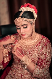 A woman dressed in traditional bridal attire adorned with intricate gold jewelry covering her neck, ears, and forehead. Her hair is styled elegantly with flowers, and her hands are decorated with henna designs. The outfit is richly embroidered with gold embellishments on a red fabric.