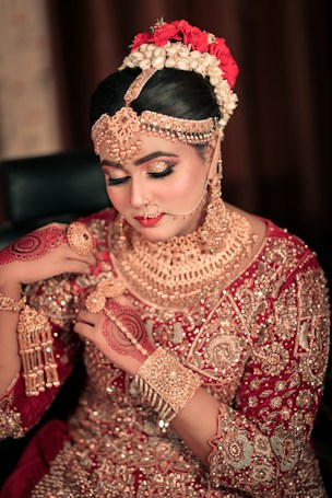 A woman dressed in traditional bridal attire adorned with intricate gold jewelry covering her neck, ears, and forehead. Her hair is styled elegantly with flowers, and her hands are decorated with henna designs. The outfit is richly embroidered with gold embellishments on a red fabric.