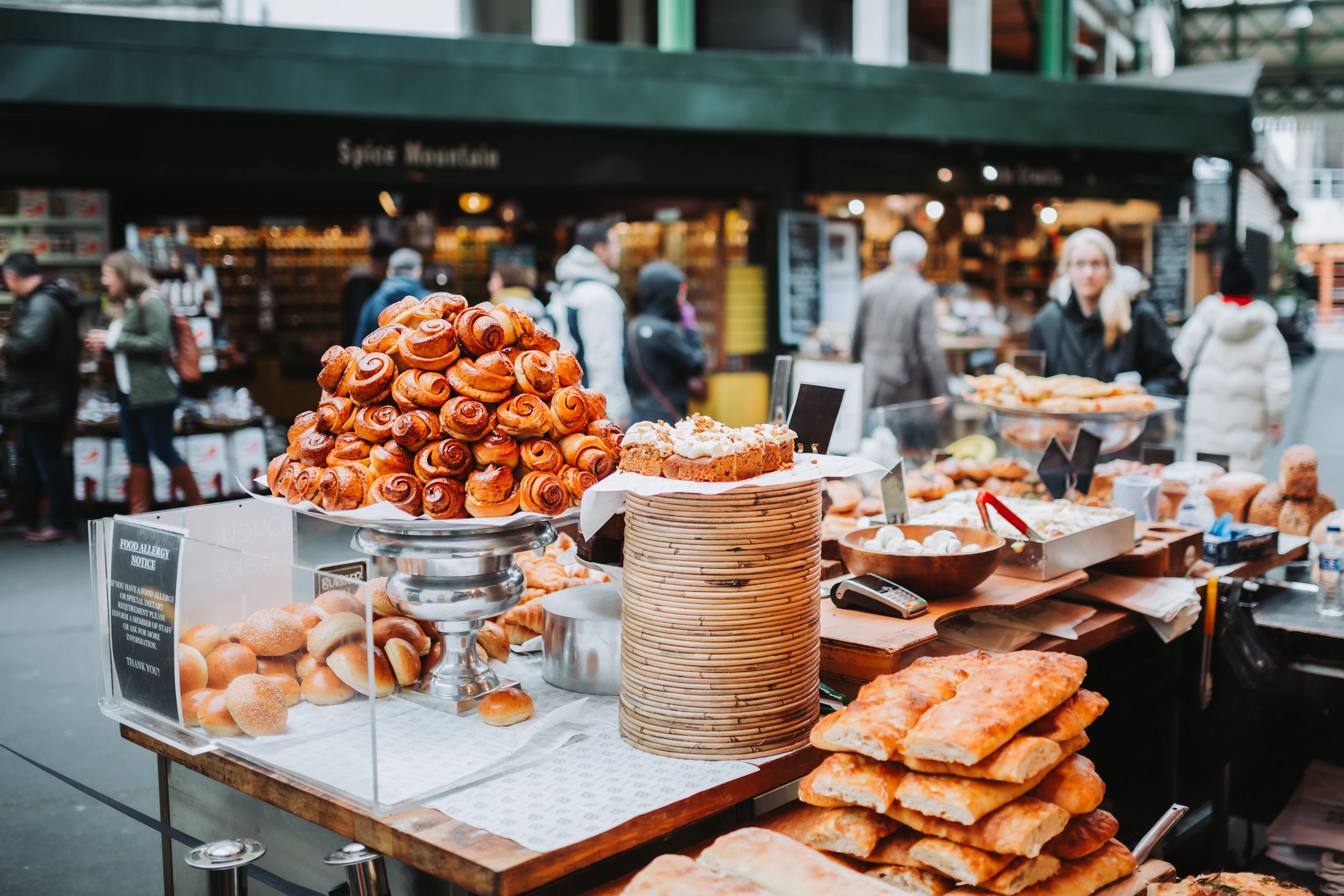 A bakery stall at Borough Market piled with buns, tarts and pastries, shoppers pressing along the aisle