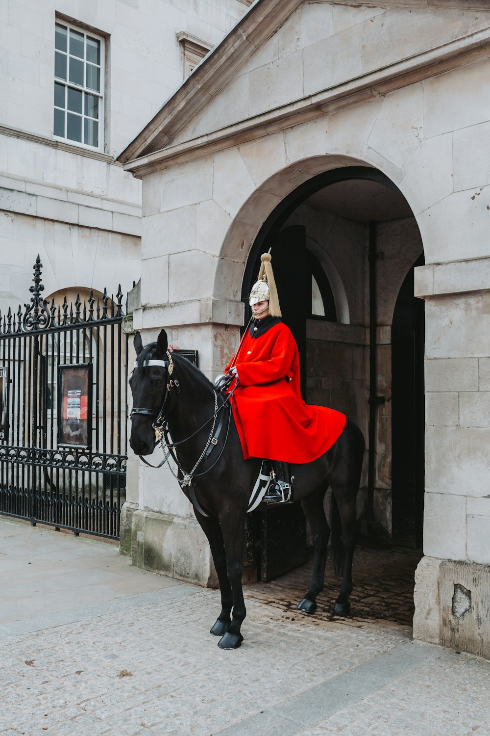 uma pessoa em um manto vermelho montando um cavalo preto com guardas de cavalo no fundo