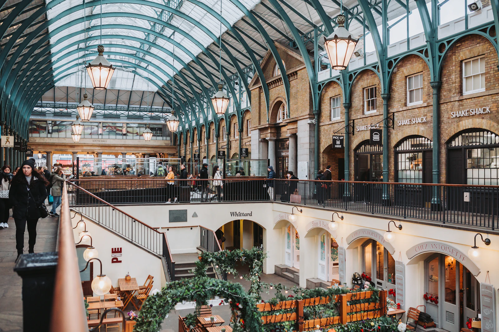 Photograph of People walk beneath the pale glass roof, green iron ribs and hanging lamps of Covent Garden market.