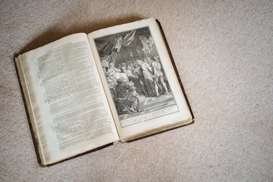 An open book is placed on a textured surface, displaying detailed text on the left page and an intricate black and white illustration on the right. The illustration features a historical or religious scene with several figures in classical attire, possibly depicting a significant event or story.