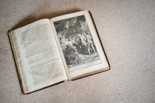 An open book is placed on a textured surface, displaying detailed text on the left page and an intricate black and white illustration on the right. The illustration features a historical or religious scene with several figures in classical attire, possibly depicting a significant event or story.