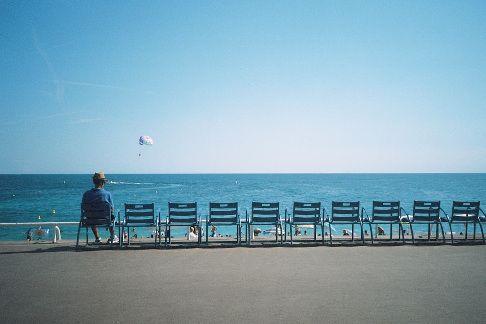 a person sitting on a bench watching a kite fly over the ocean
