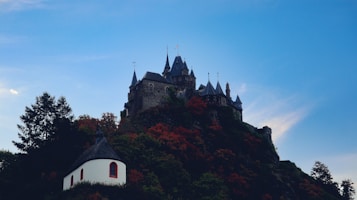 A picturesque castle sits atop a hill surrounded by vibrant autumn foliage. The castle's pointed towers and stone walls give it a fairytale-like appearance. Below it, a small white building with a dark roof is nestled among the trees. The sky is clear with a gentle gradient of blue.