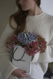 A person wearing a cozy white knit sweater holds a white shopping bag filled with hydrangeas in shades of pink and purple. The shopping bag has the brand logo 'Chanel' printed on it. The background is soft and neutral, emphasizing the flowers and the sweater.