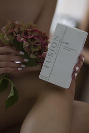 A person holds a hydrangea flower in one hand and a box labeled 'Fusion Meso Therapy F-PPC' in the other. The focus is on the product and flower, with soft lighting and muted background.