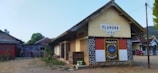 A rustic building with a sign reading 'SLAHUNG +154' stands in a rural area. The structure has a tiled roof and walls that combine brick and painted sections, some of which feature a circular emblem. Surrounding the building are other small structures and elements like potted plants and stacks of wooden pallets. Trees and hills are visible in the background under a clear sky.