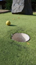 A close-up view of a mini-golf course with a yellow golf ball positioned near a hole. The surface is covered with green artificial turf and a large rock is visible in the background.