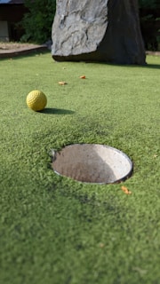 A close-up view of a mini-golf course with a yellow golf ball positioned near a hole. The surface is covered with green artificial turf and a large rock is visible in the background.