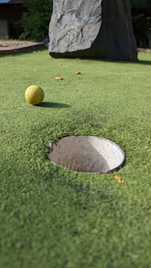 A close-up view of a mini-golf course with a yellow golf ball positioned near a hole. The surface is covered with green artificial turf and a large rock is visible in the background.