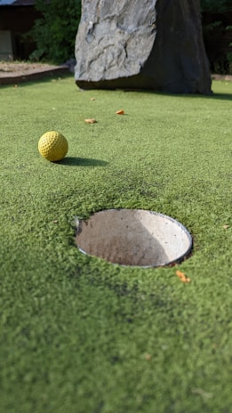 A close-up view of a mini-golf course with a yellow golf ball positioned near a hole. The surface is covered with green artificial turf and a large rock is visible in the background.