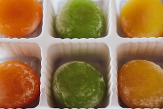 Six colorful round mochi pieces are arranged in a white plastic tray, featuring two each of orange, green, and yellow. The sweets have a slightly glistening surface, suggesting they are fresh or slightly sticky.