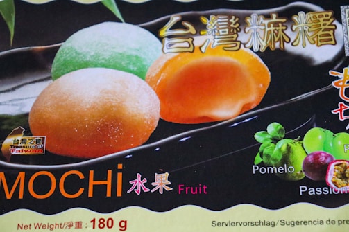 A package featuring colorful mochi, a Japanese dessert, with three visible mochi balls in pastel green, orange, and pale yellow. The background includes a label indicating 'Treasure of Taiwan' and text in multiple languages including English and Chinese. The package highlights fruit flavors with images of pomelo and passion fruit.