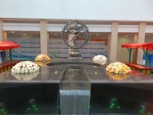 A bronze statue depicting a dancing figure is centrally displayed on a reflective black platform. Surrounding the statue, there are four floral arrangements featuring a variety of colorful flowers, including whites, reds, and yellows. The background includes a wall with logos and branding, and there are red-roofed, green-framed stalls on either side.