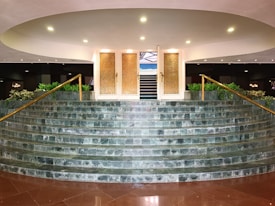 A grand staircase made of green marble tiles leads up to a set of ornate golden doors with textured glass panels. The staircase is bordered by golden railings and flanked by potted plants, creating a sense of elegance and luxury. Soft, ambient lighting highlights the entrance, adding a warm and inviting atmosphere.