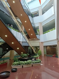 A multi-level indoor space featuring modern escalators and numerous indoor plants arranged in planters along the base. The floors are polished with a glossy finish and the environment is well-lit, suggesting a commercial or office building setting with open atrium-like architecture.