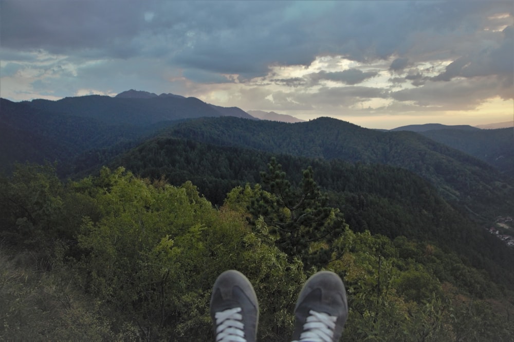 a person's legs and feet on a hill with trees and mountains in the background