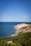 A scenic coastline with a clear blue sky and a vast expanse of blue ocean meeting a rocky shoreline. Lush green vegetation covers the cliff tops, leading down to sandy beaches and rugged cliffs.