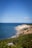 A scenic coastline with a clear blue sky and a vast expanse of blue ocean meeting a rocky shoreline. Lush green vegetation covers the cliff tops, leading down to sandy beaches and rugged cliffs.