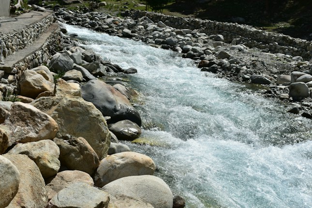 A clear mountain stream flows rapidly over rocky terrain, bordered by large, smooth stones. The water is sparkling and appears cool, cascading over the rocks with a slight white froth. On one side, a stone pathway runs parallel to the stream, lined with more rocks.